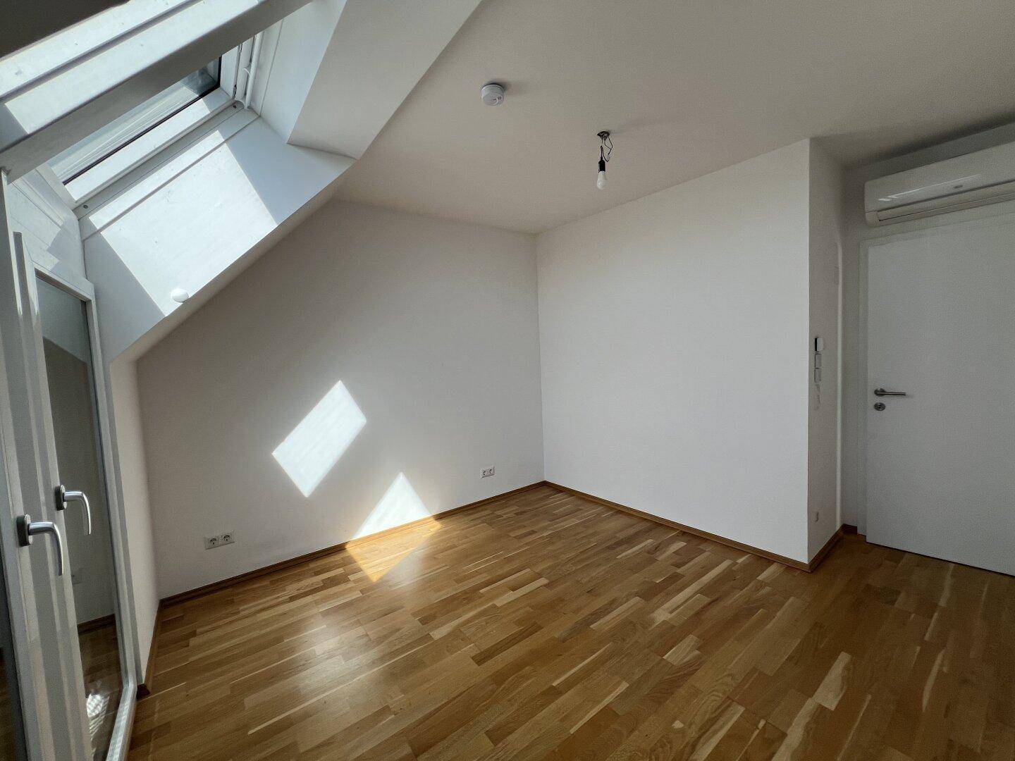 Helles Zimmer mit Holzboden, Dachfenster und weiß gestrichenen Wänden.