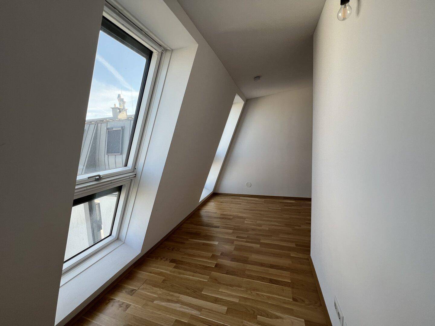 Helles Zimmer mit Holzboden und schrägen Dachfenstern, die einen Blick nach draußen bieten.