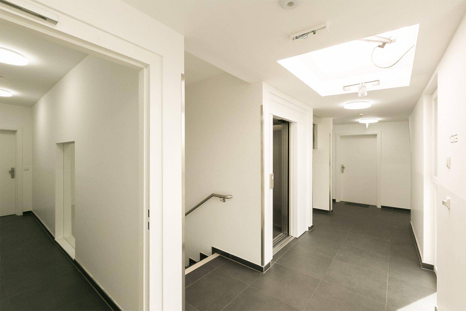 Heller Flur im Gebäude mit Aufzug, Treppe und Fliesenboden.