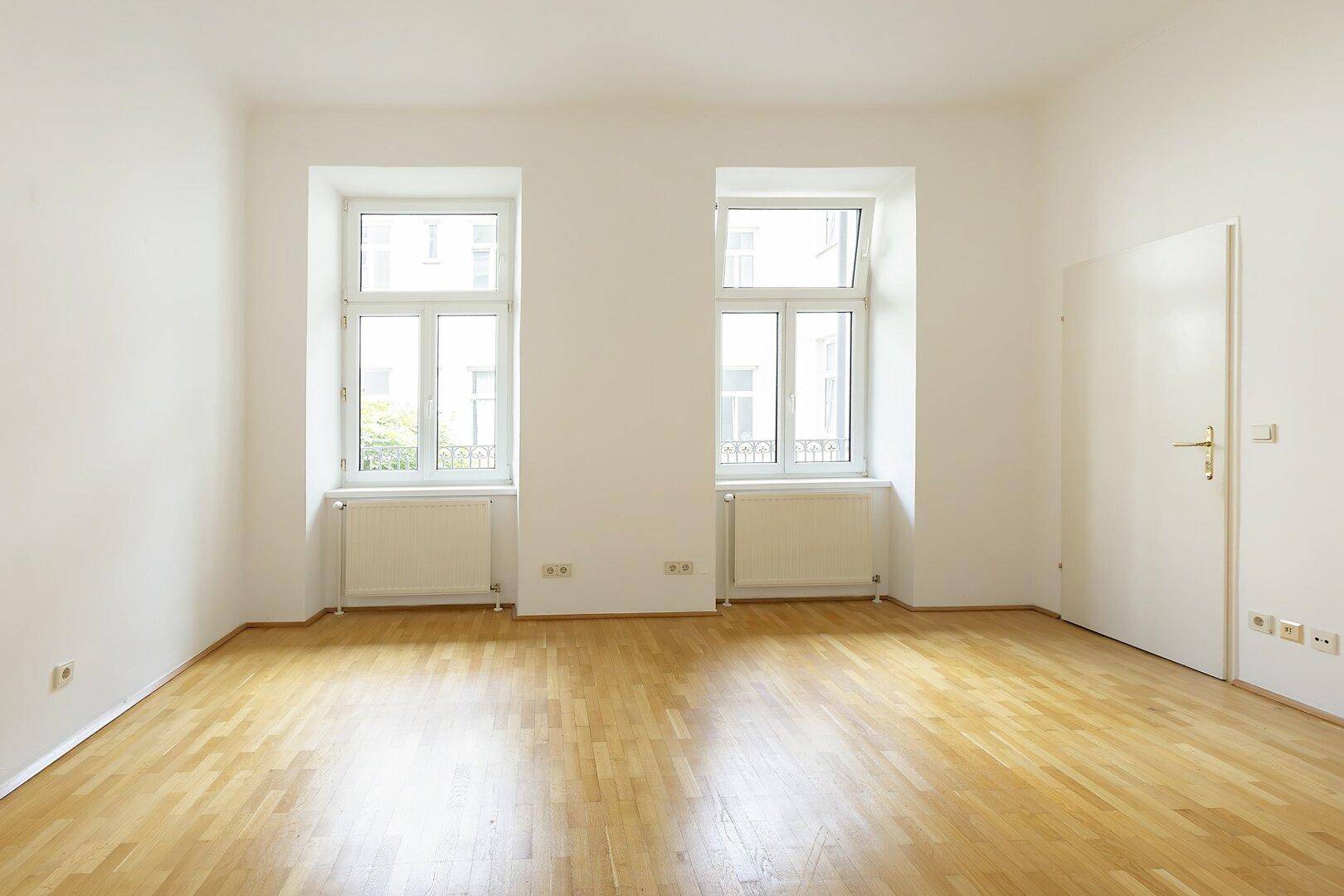 Helles, leeres Zimmer mit zwei Fenstern, Parkettboden und weißen Wänden.