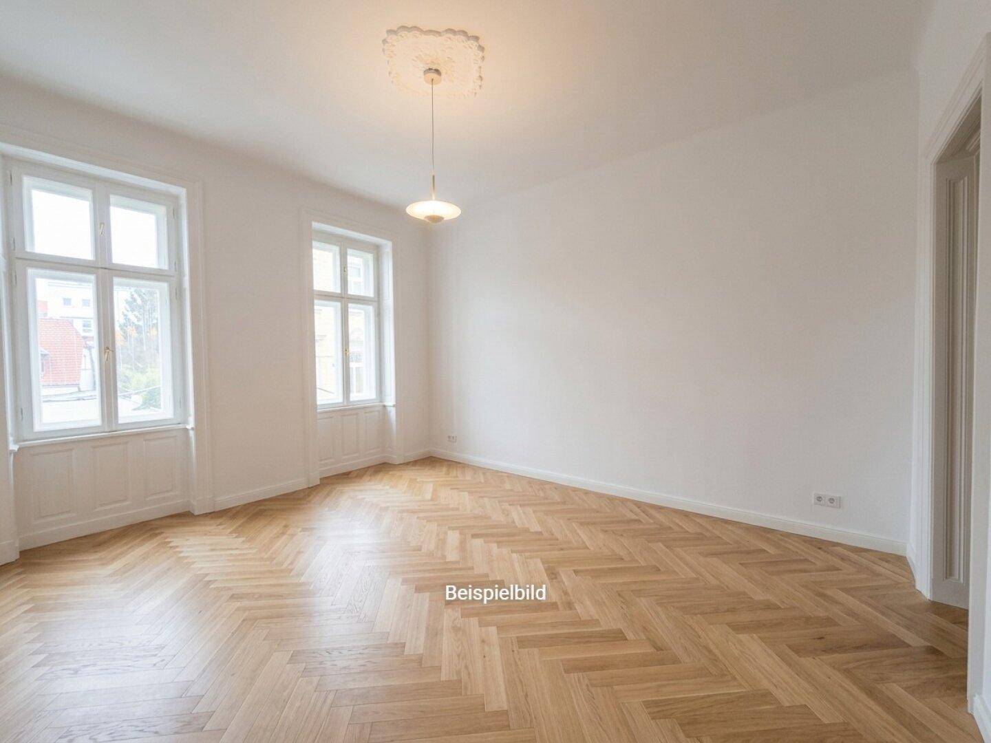Helles Zimmer mit Fischgrätparkett, hohen Decken und großen Fenstern, die viel Tageslicht hereinlassen.