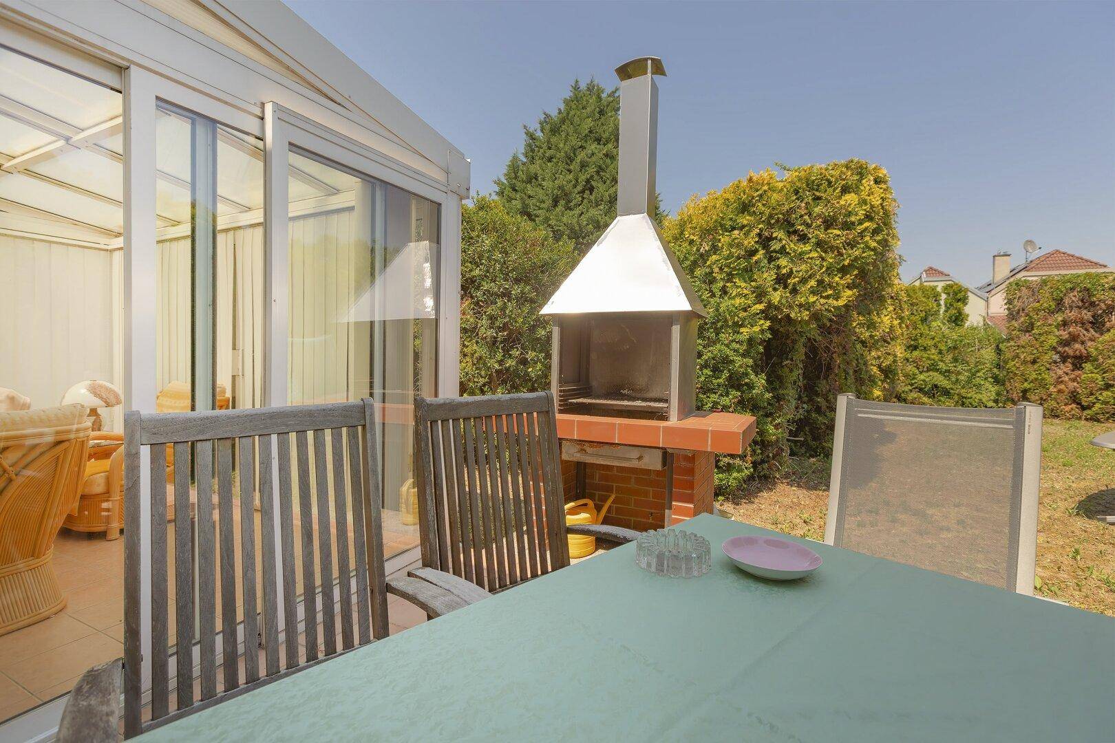 Sonniger Terrassenbereich mit Esstisch, Stühlen und gemauertem Grill, angrenzend an den Wintergarten.
