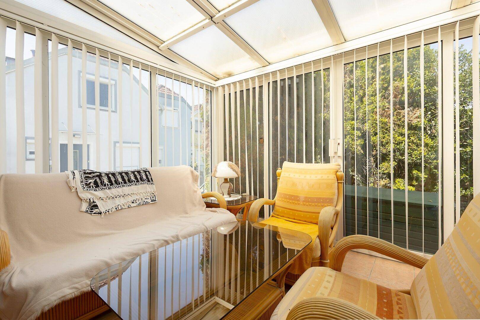 Heller Wintergarten mit Rattanmöbeln und großen Fenstern, die viel Tageslicht hereinlassen.