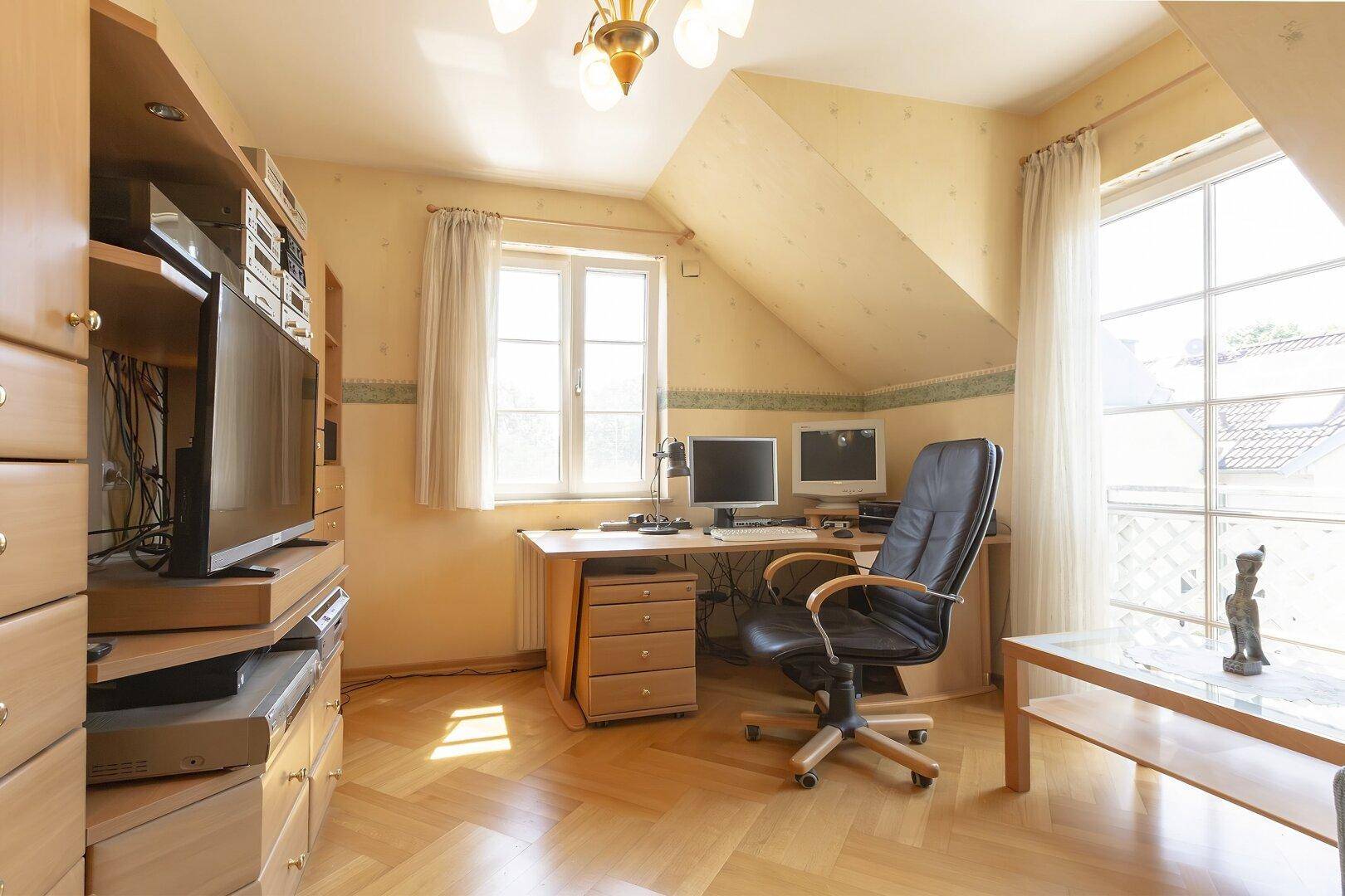 Helles Arbeitszimmer im Dachgeschoss mit Schreibtisch, Computern und Parkettboden, mit großen Fenstern.