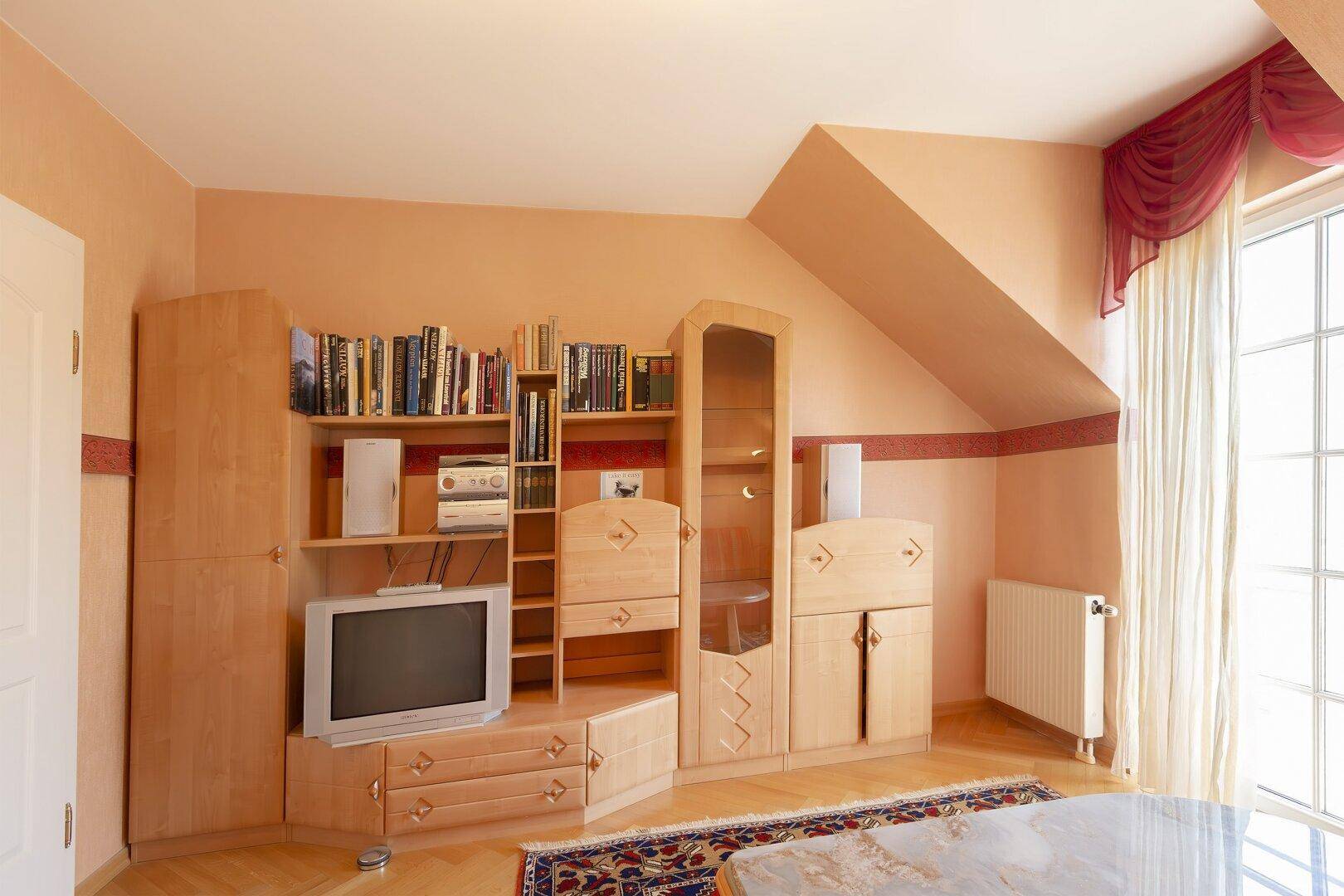 Schlafzimmer im Dachgeschoss mit schräger Decke, Holzmöbeln und Fernseher, hell und funktional.
