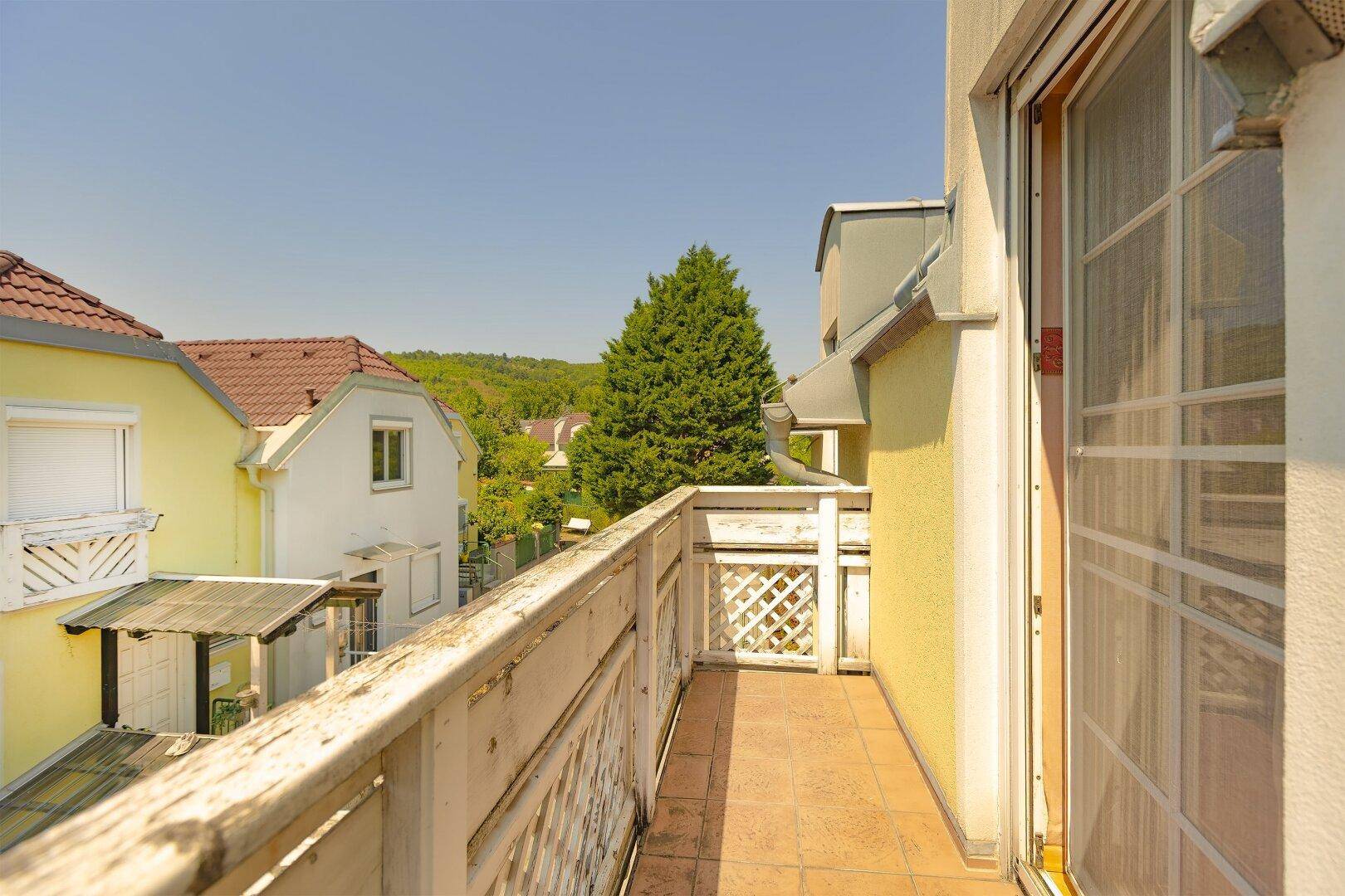 Langer Balkon mit Holzgeländer und Blick auf die grüne Umgebung und Nachbarhäuser.