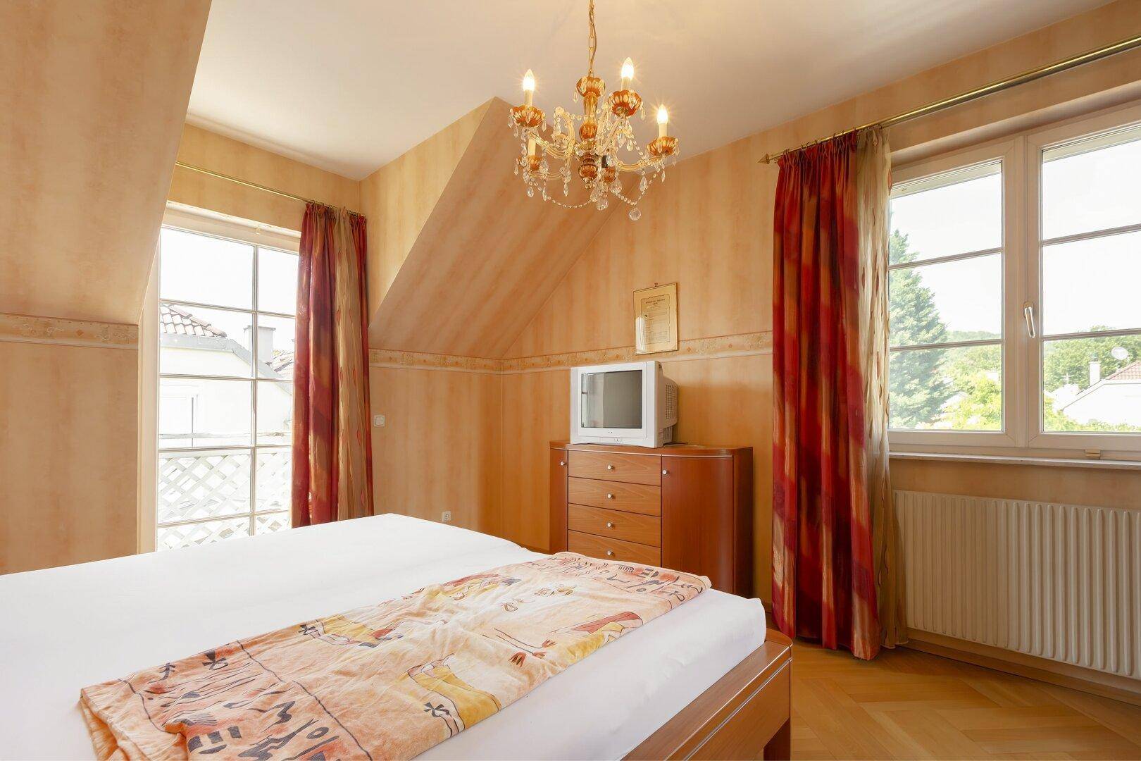 Gemütliches Schlafzimmer mit Dachschräge, großen Fenstern und traditionellem Kronleuchter.