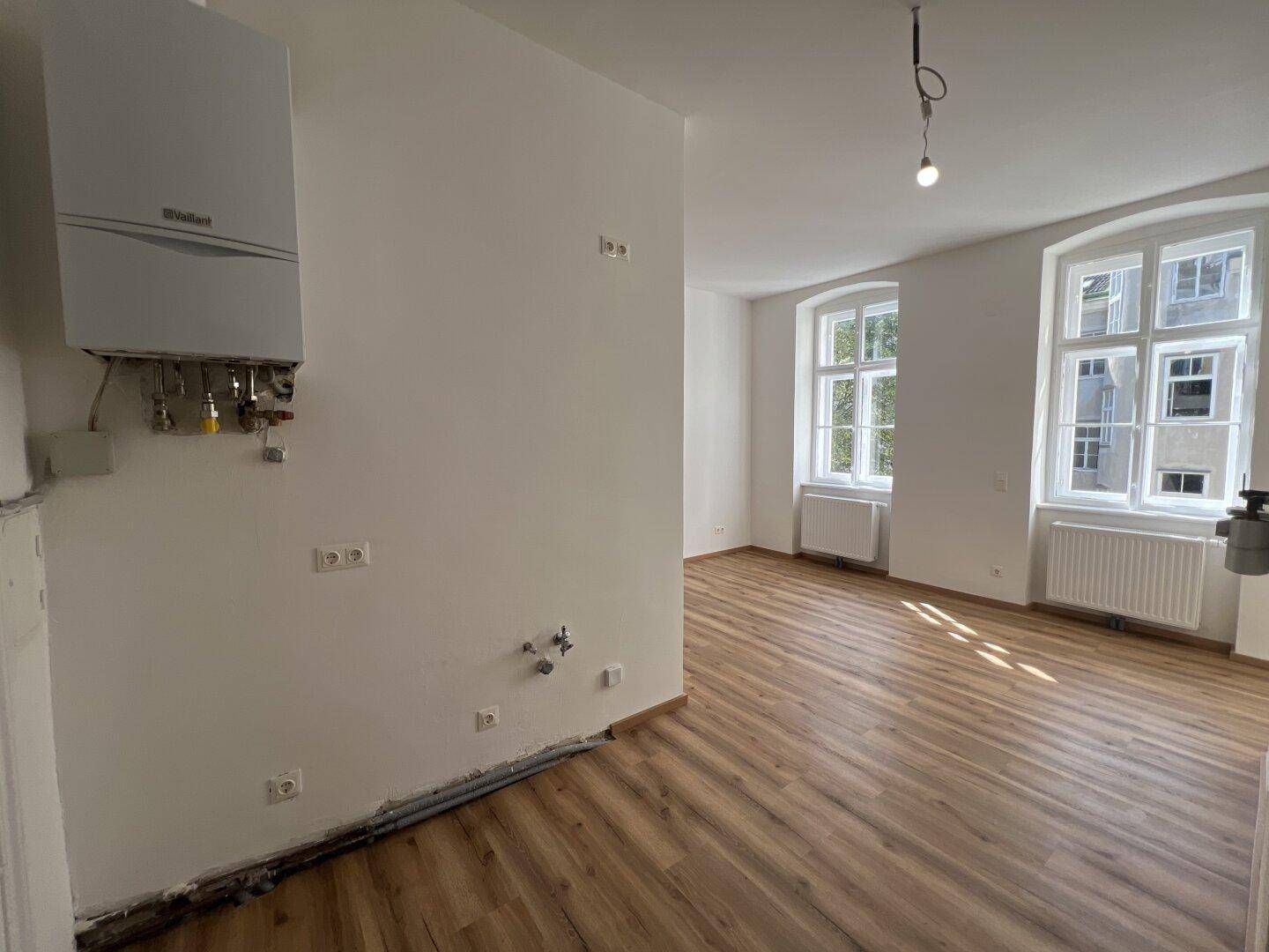Raum mit Holzlaminatboden, Gastherme und Anschlüssen für eine Küche, hell durch große Fenster.