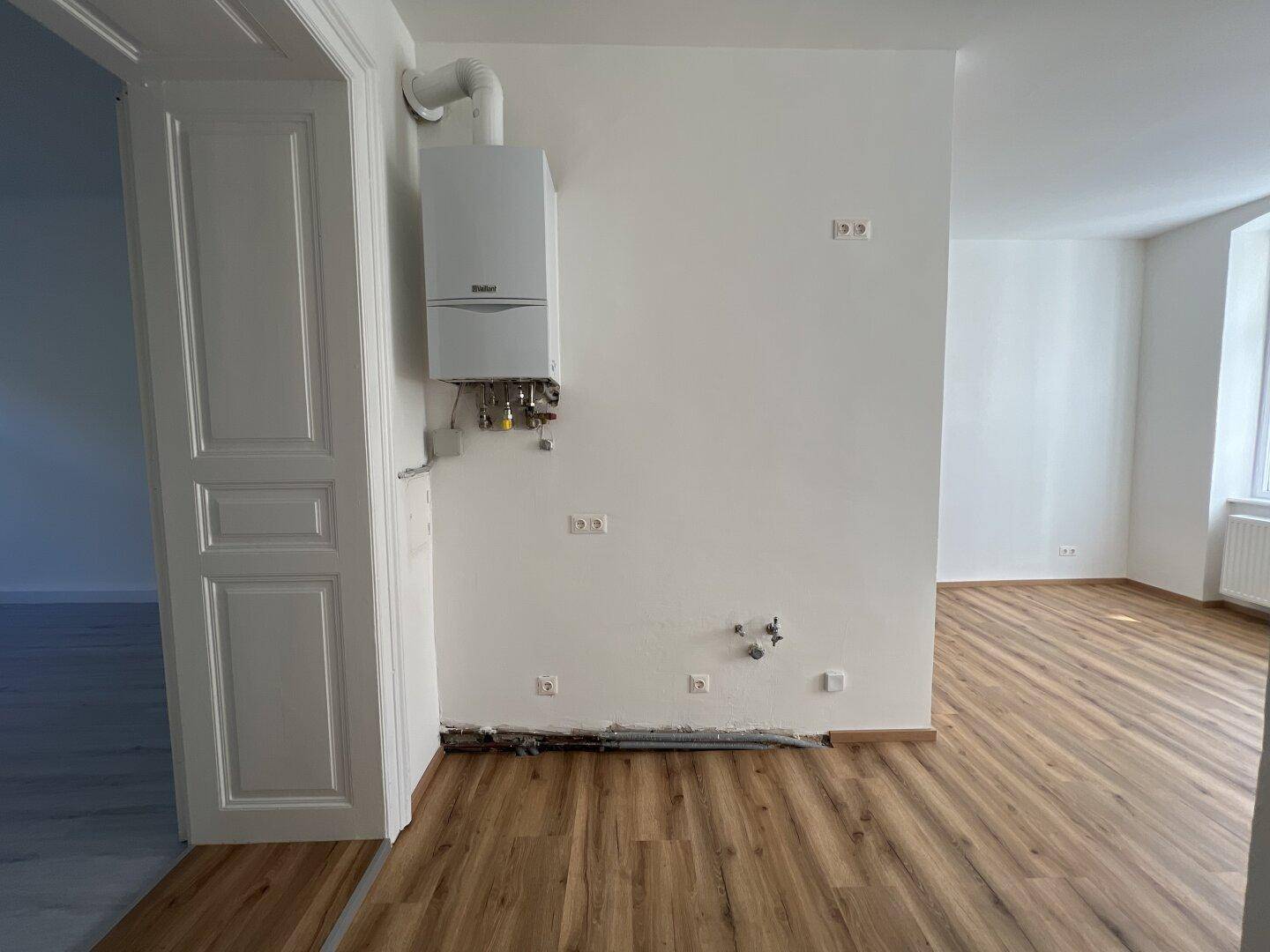 Bereich mit Gastherme und Küchenanschlüssen, angrenzend an einen hellen Raum mit Holzlaminatboden.