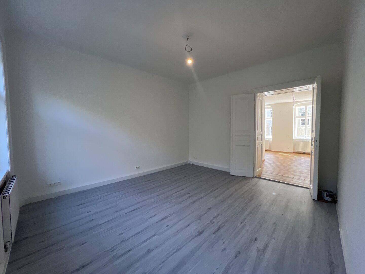 Geräumiges, leeres Zimmer mit grauem Laminatboden und einer Doppeltür zu einem weiteren Raum.