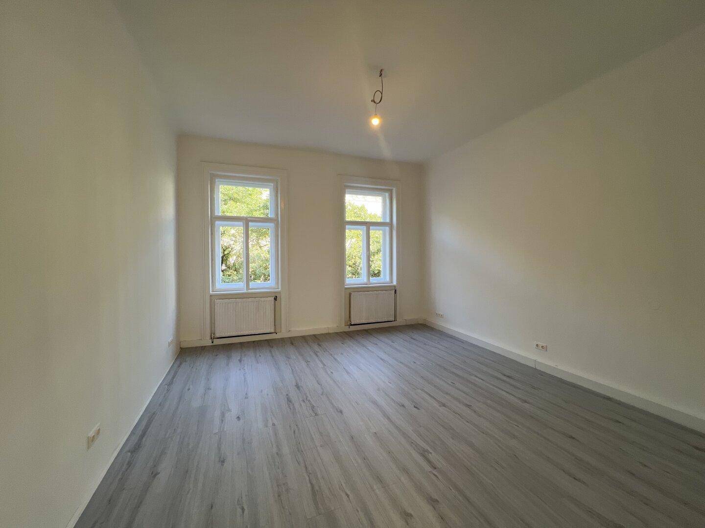 Heller, leerer Raum mit grauem Laminatboden und zwei Fenstern, die viel Sonnenlicht hereinlassen.