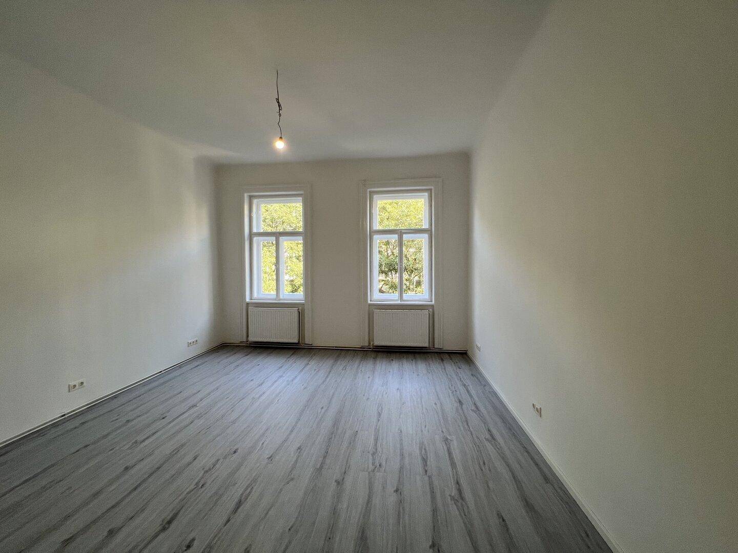 Leeres Zimmer mit grauem Laminatboden und zwei Fenstern, die einen Blick ins Grüne bieten.