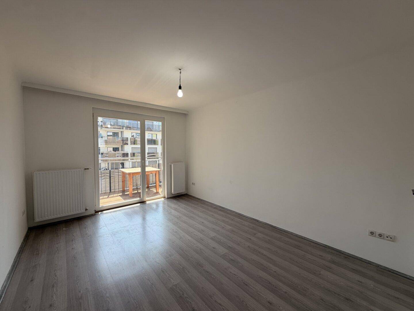 Heller, leerer Raum mit grauem Laminatboden, weißen Wänden und Zugang zum Balkon.