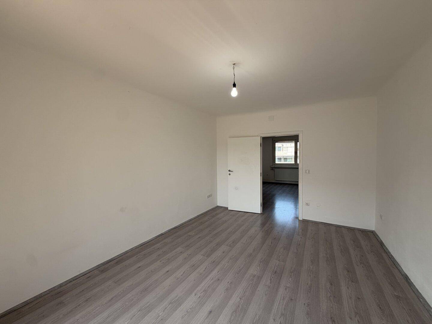 Geräumiger, leerer Raum mit grauem Laminatboden und weiß gestrichenen Wänden.