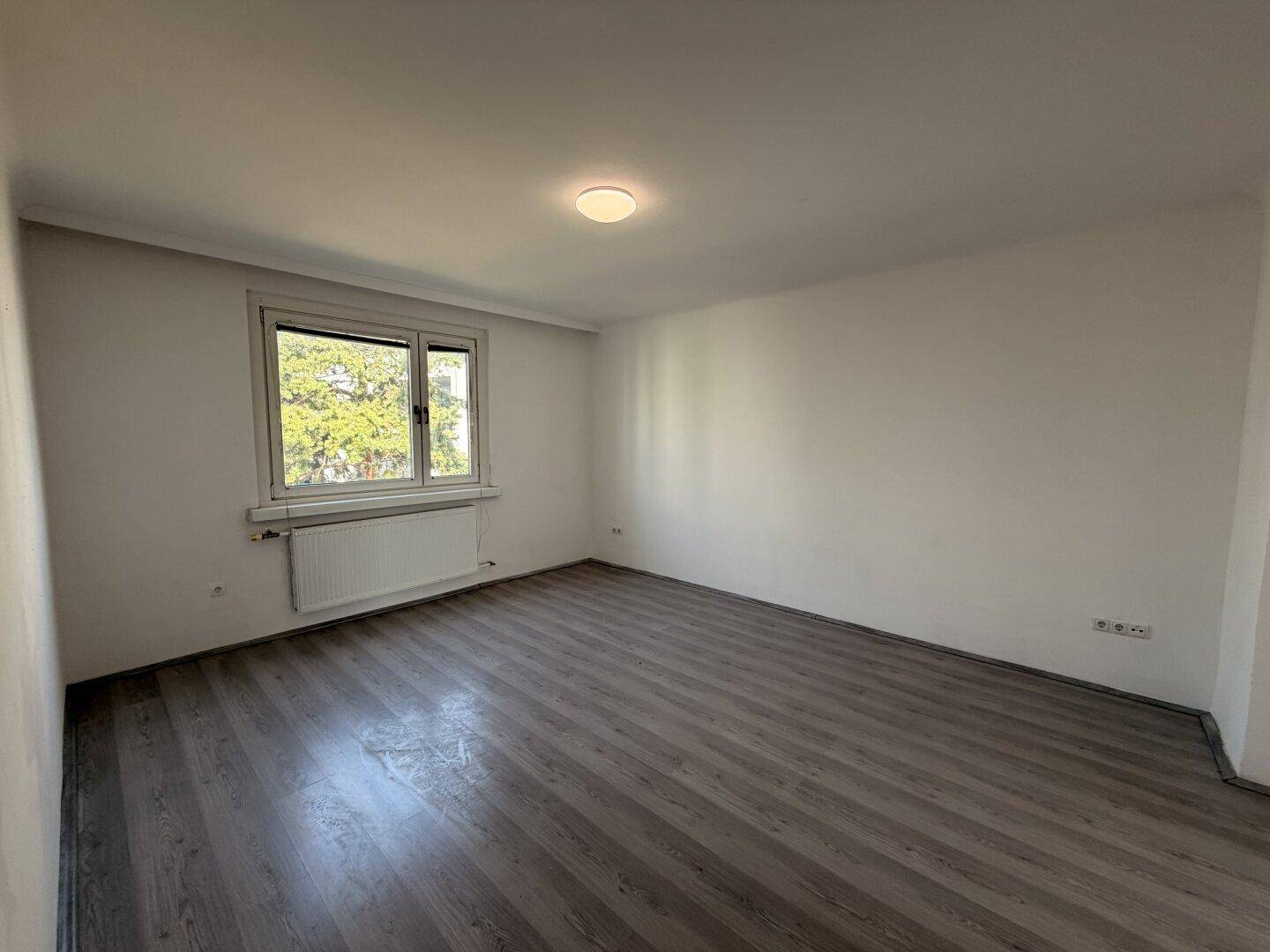 Leeres Schlafzimmer mit grauem Laminatboden, weißen Wänden und Fenster mit Blick ins Grüne.