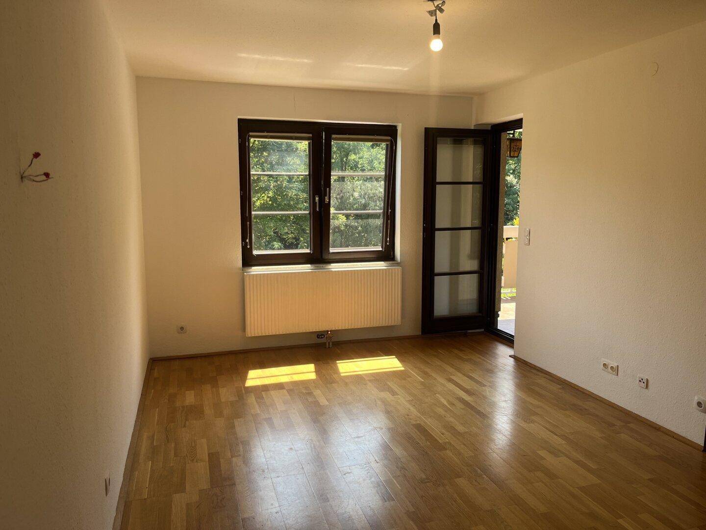 Helles, leeres Zimmer mit Parkettboden, großem Fenster und Zugang zu einem Balkon.