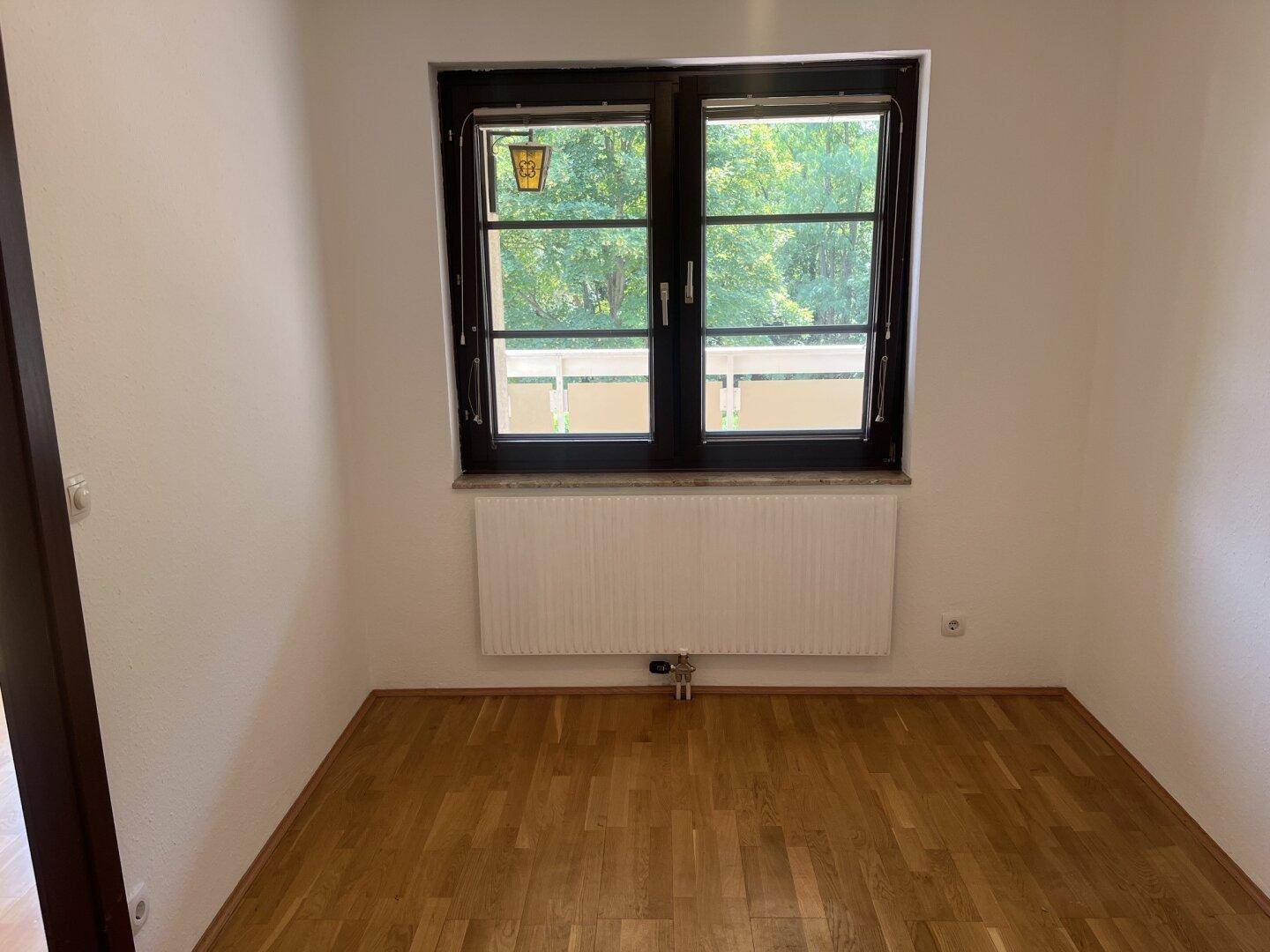 Leeres Zimmer mit Parkettboden, großem Fenster und Heizkörper unter der Fensterbank.