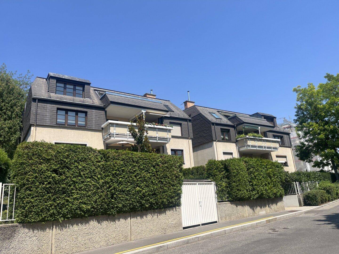 Mehrfamilienhaus mit Mansarddach, Balkonen und gepflegter Hecke an der Straße.