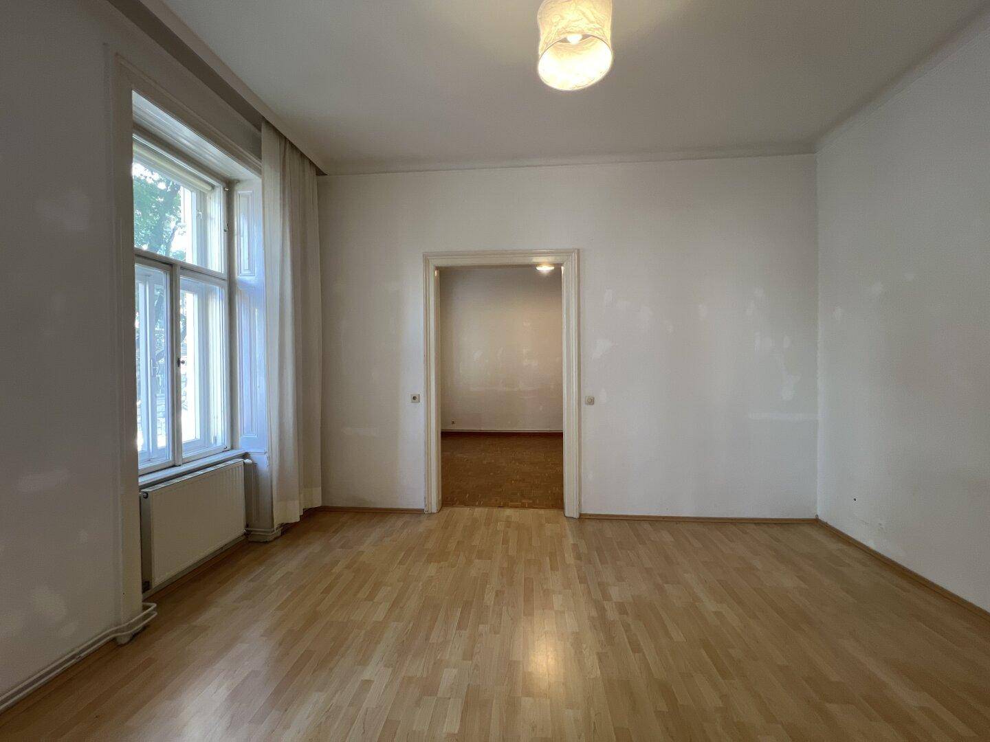 Geräumiges Wohnzimmer mit Holzboden und viel Tageslicht durch ein großes Fenster.
