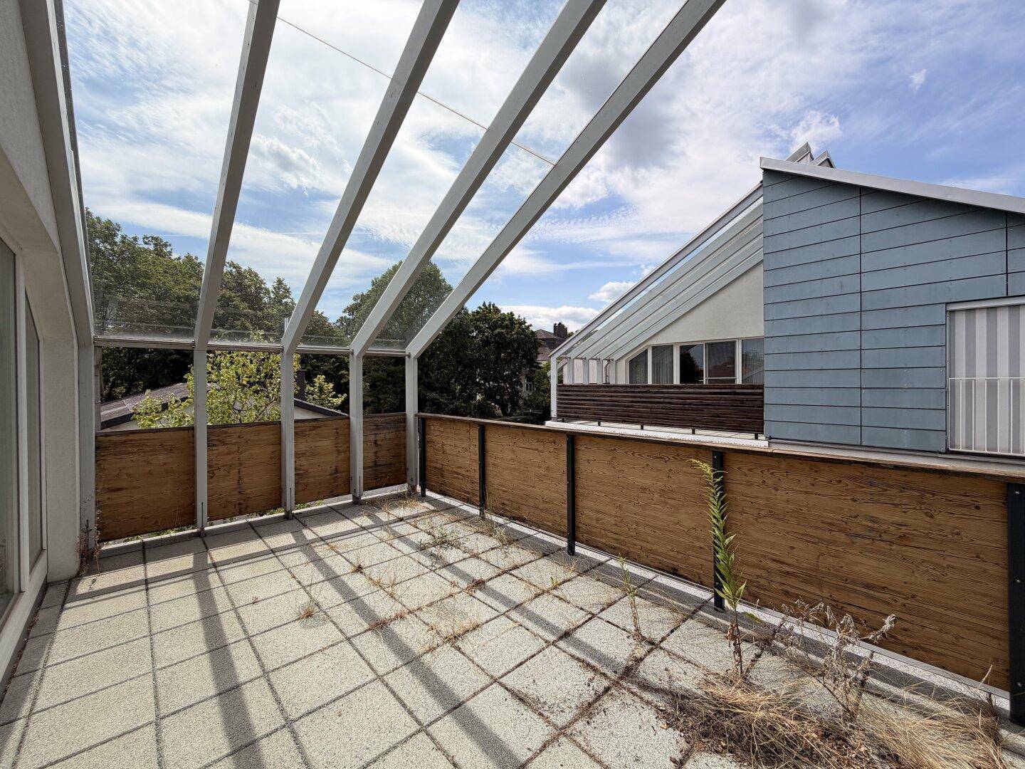 Geräumige Terrasse mit Fliesenboden und einer transparenten Überdachung, umgeben von Bäumen.