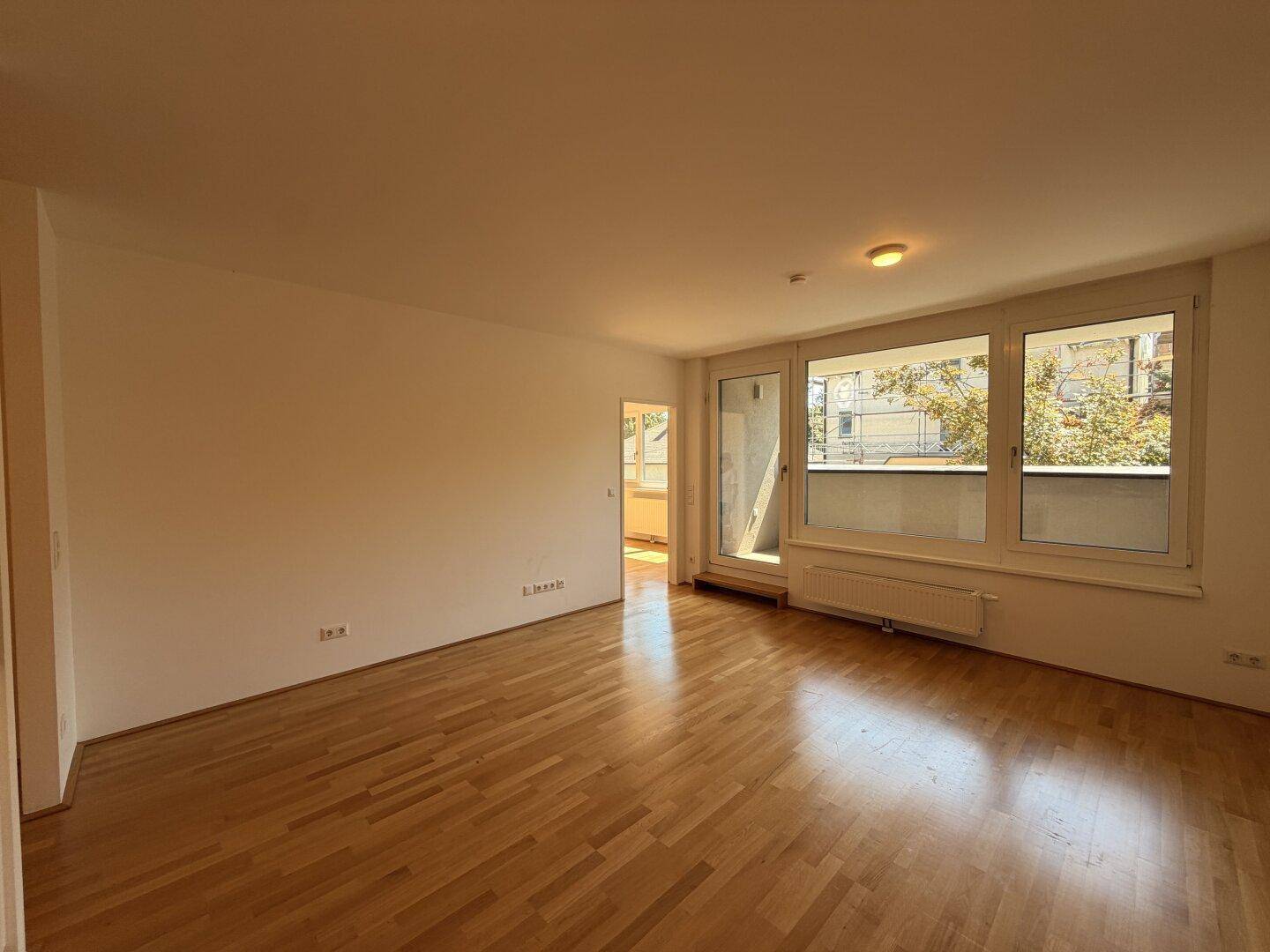 Geräumiges Wohnzimmer mit hellem Parkettboden, großen Fenstern und Zugang zum Balkon.
