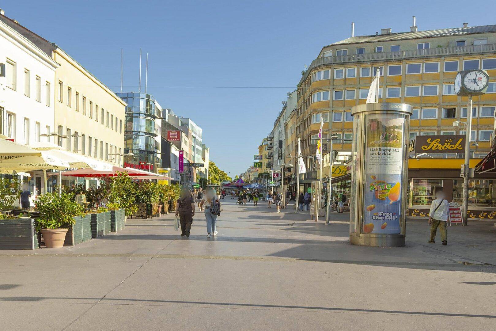 Belebte Fußgängerzone mit Geschäften, Restaurants und Passanten unter klarem blauem Himmel.