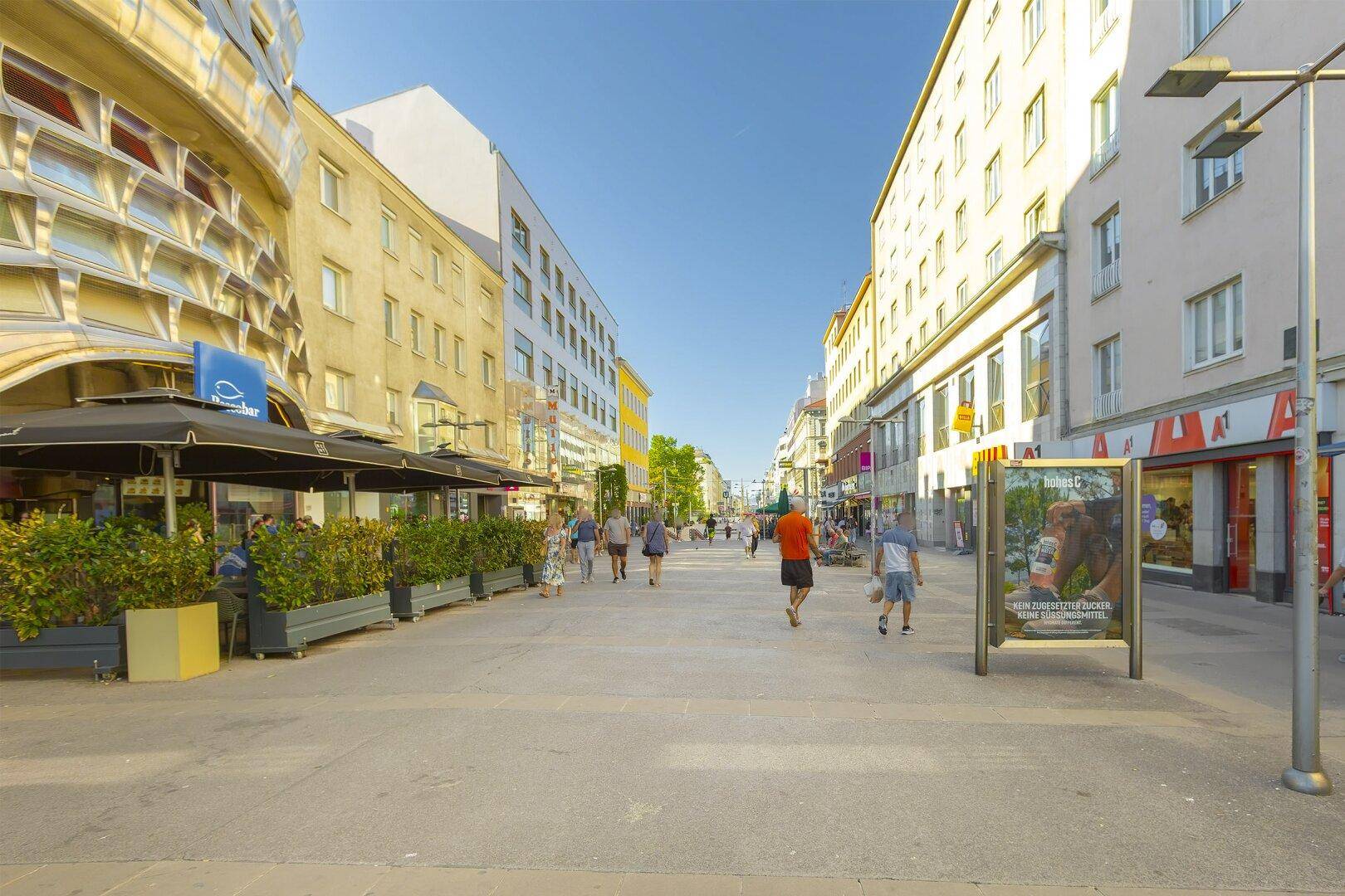 Weitläufige Fußgängerzone mit zahlreichen Geschäften und Cafés, belebt von Passanten an einem sonnigen Tag.