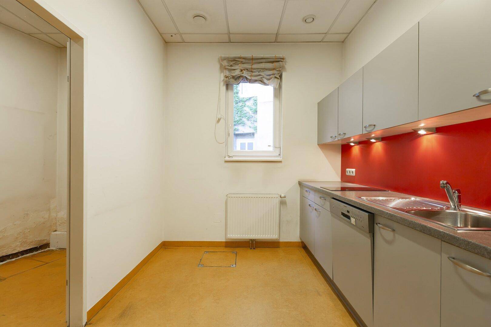 Küchenbereich mit Fenster, grauen Unterschränken, roter Rückwand und gelbem Linoleumboden.