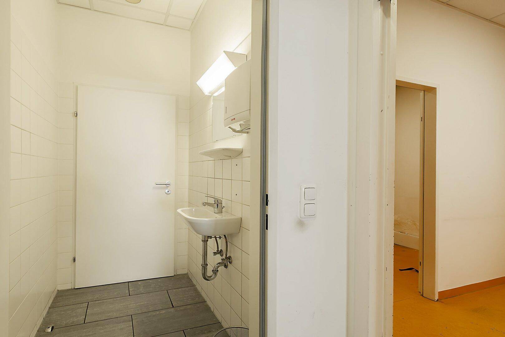 Kleiner Toilettenraum mit Waschbecken, weißen Fliesen an den Wänden und dunklem Fliesenboden.