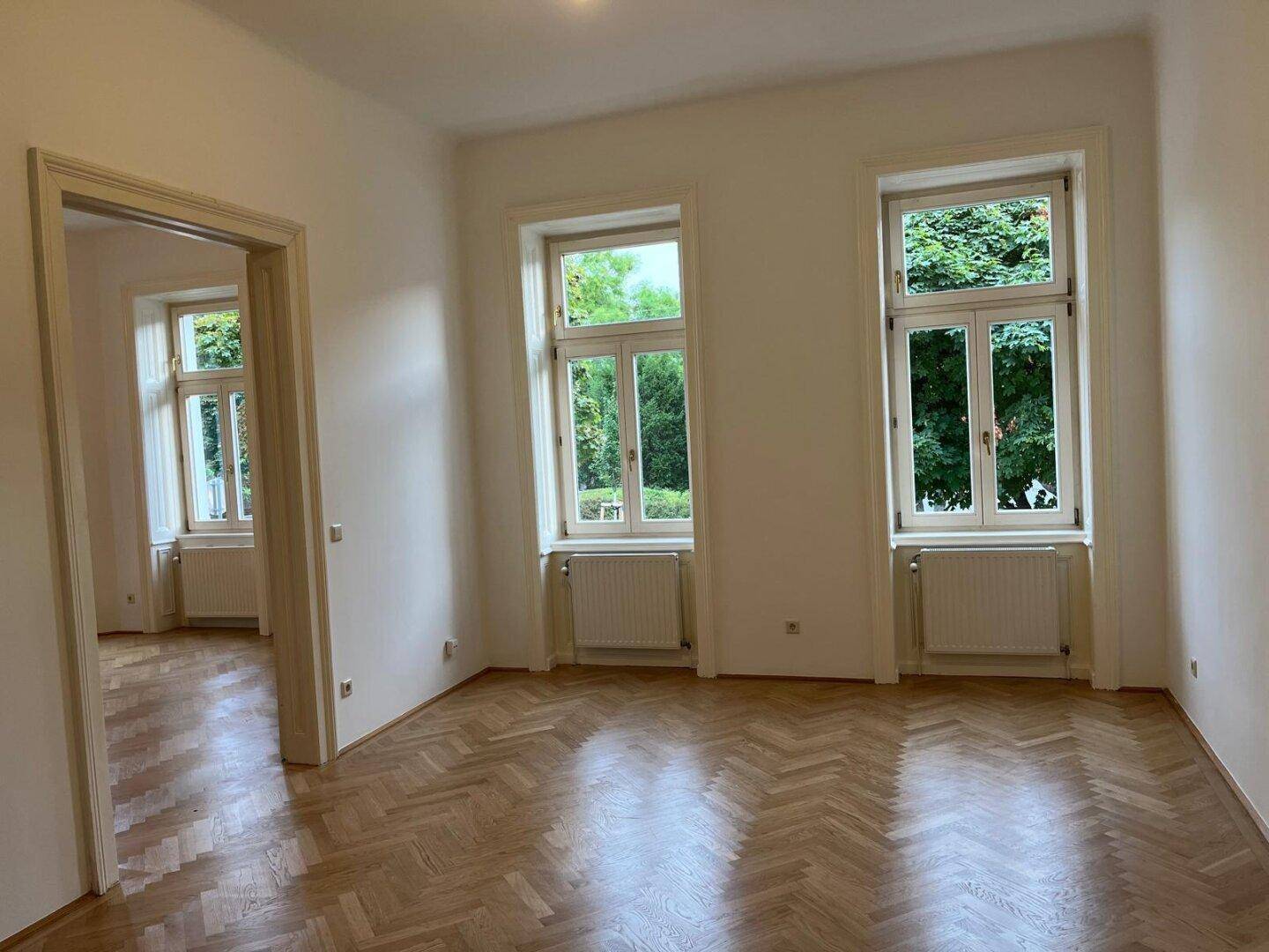 Heller Raum mit Fischgrätparkett und zwei Fenstern, verbunden mit einem weiteren Zimmer.