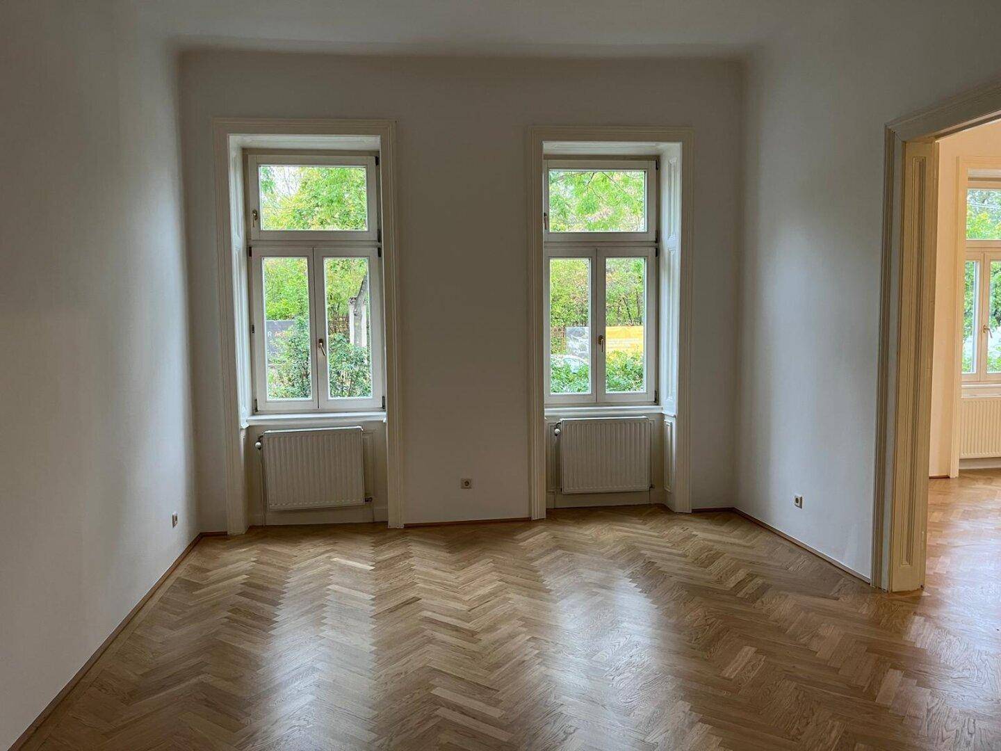 Freundlicher Raum mit Fischgrätparkett und zwei großen Fenstern mit Blick ins Grüne.