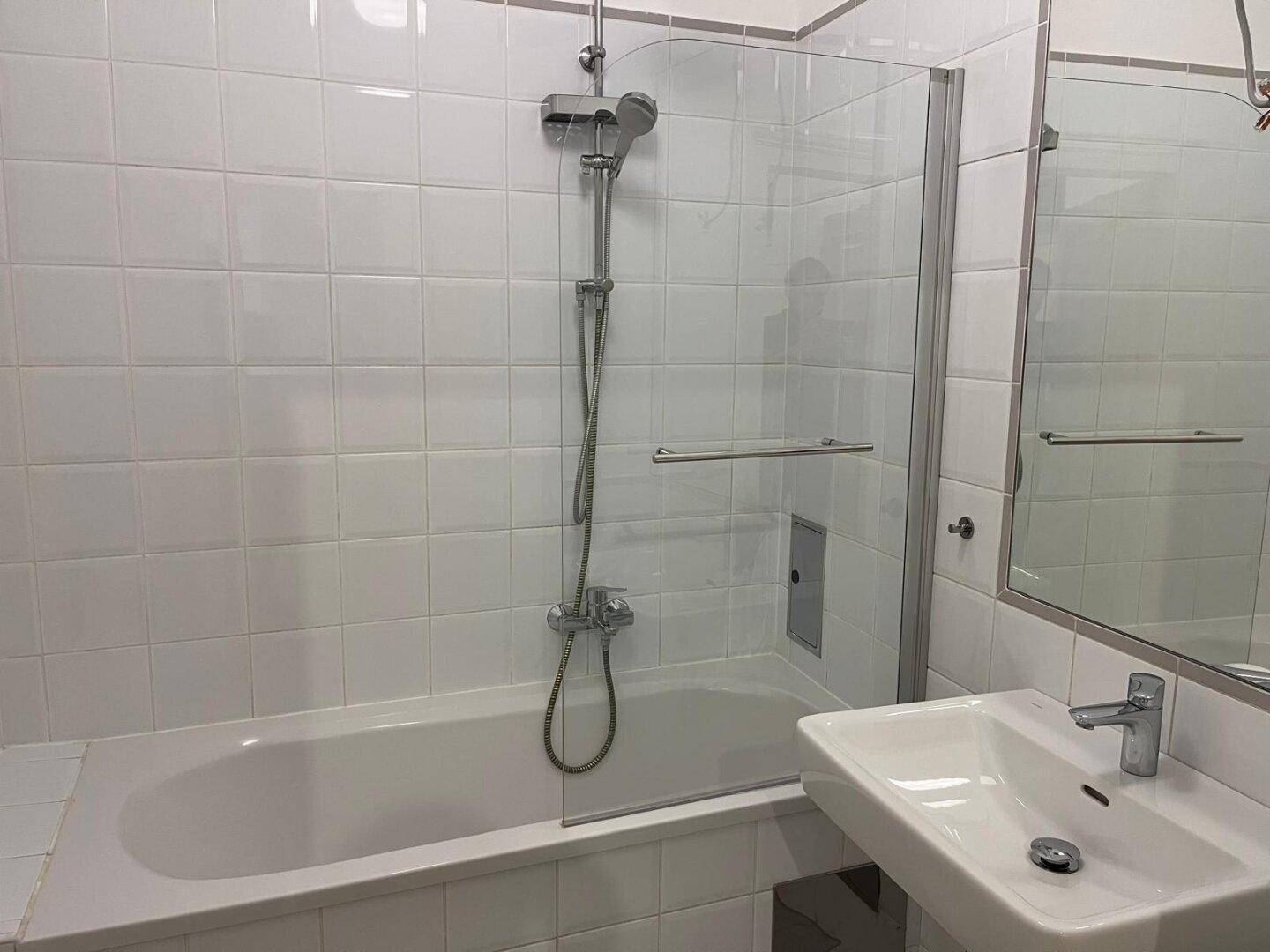 Helles Badezimmer mit Badewanne, Duschvorrichtung und weiß gefliesten Wänden.