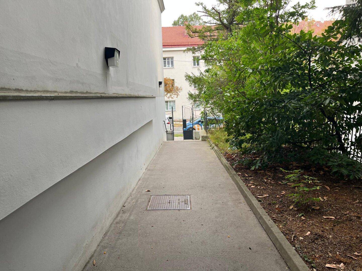 Schmaler Weg entlang der Gebäudeseite mit Bäumen und Sträuchern.
