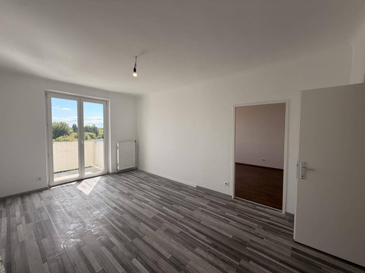 Helles, leeres Wohnzimmer mit Laminatboden, weißen Wänden und Zugang zu einem Balkon.