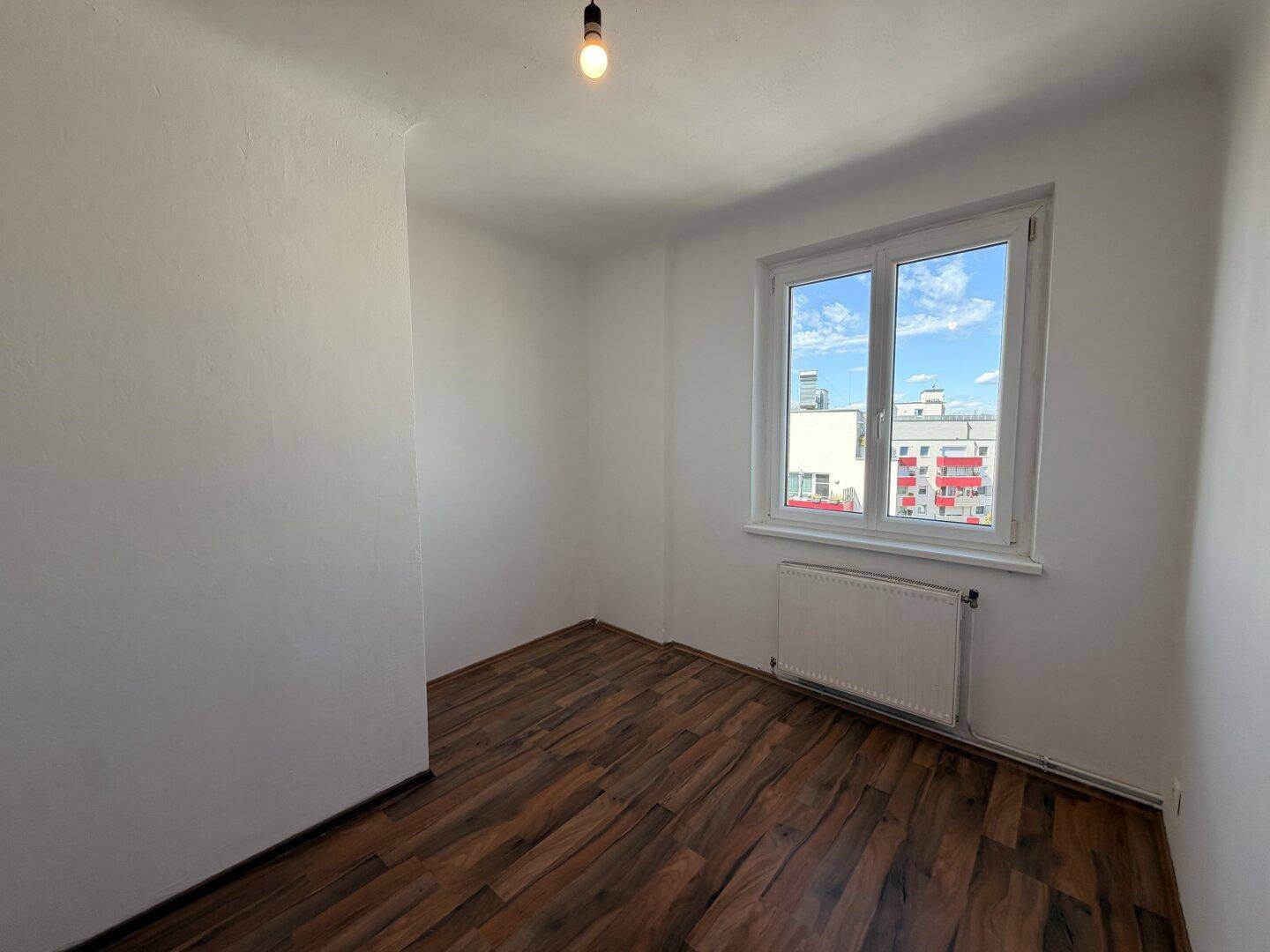 Kleineres, helles Zimmer mit dunklem Laminatboden, weißen Wänden und einem Fenster mit Stadtblick.