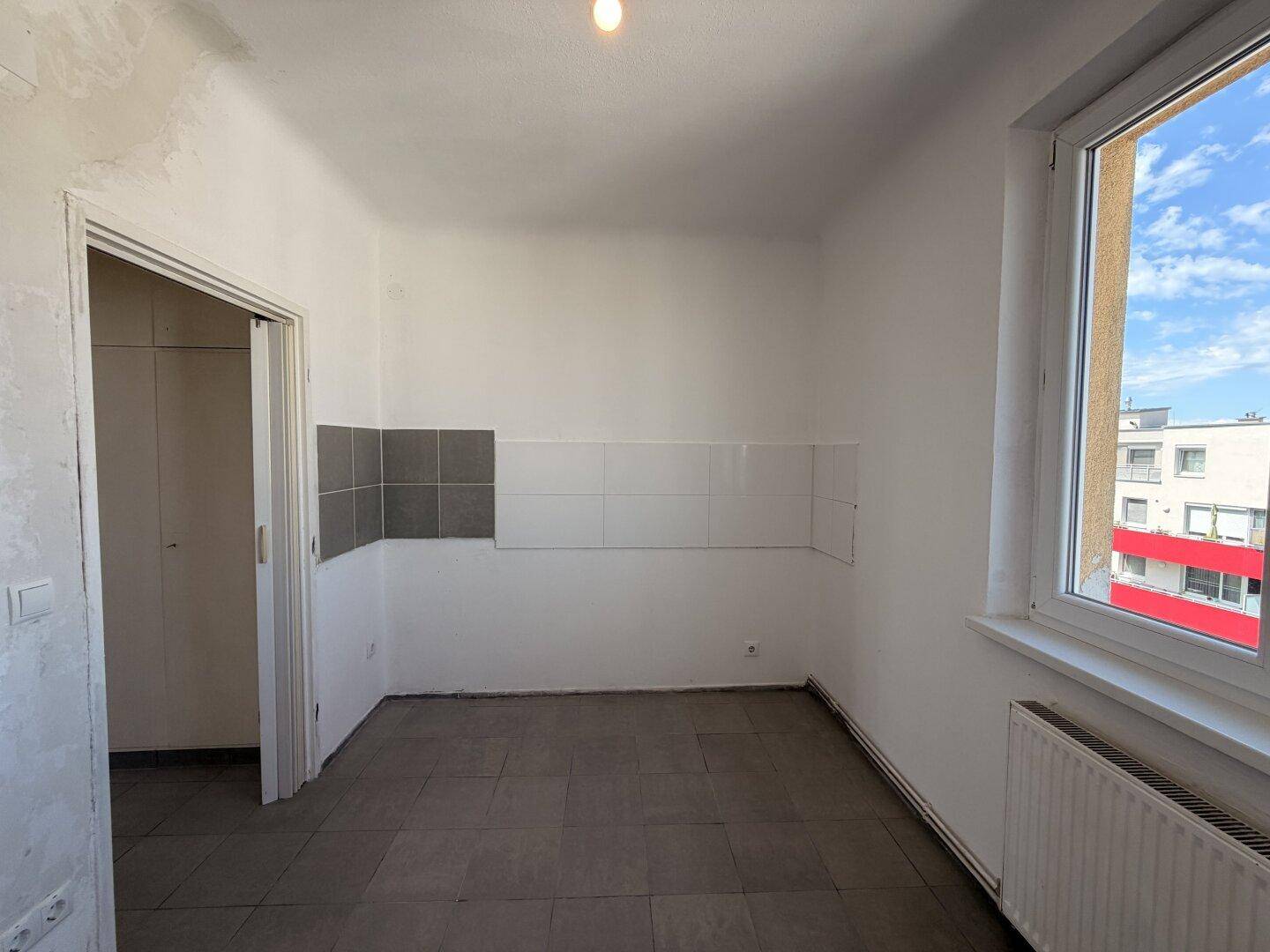 Leere Küche mit grauen Fliesen, teilweise gefliesten Wänden und einem Fenster für Tageslicht.