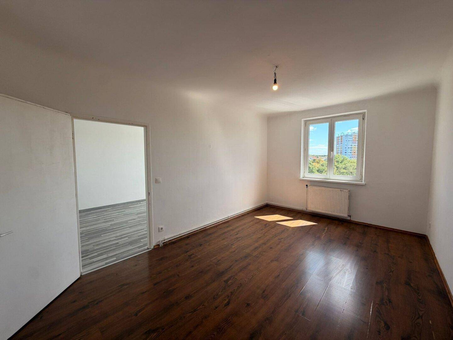 Geräumiges, leeres Zimmer mit dunklem Laminatboden, weiß gestrichenen Wänden und einem Fenster.