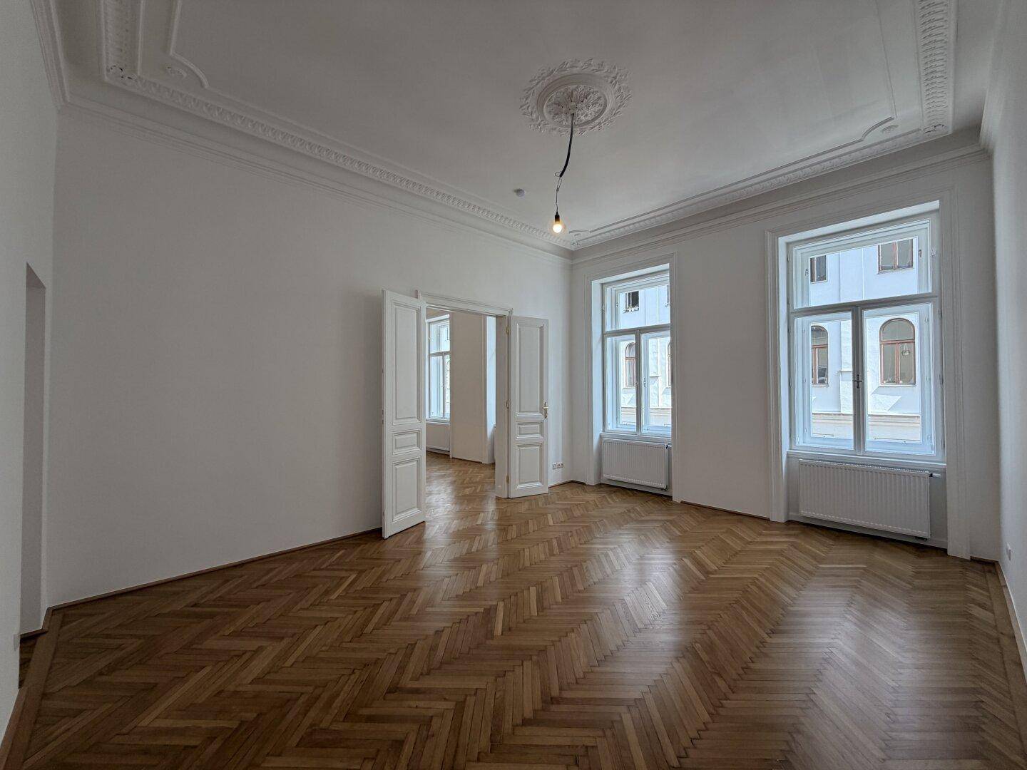 Helles Wohnzimmer mit Fischgrätparkett, hohen Decken und aufwendigem Stuck. Große Fenster lassen viel Tageslicht herein.
