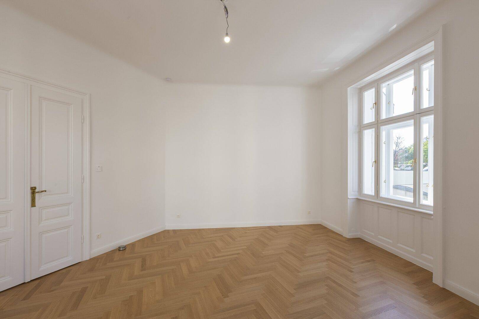Geräumiges Zimmer mit Fischgrätparkett und einem großen Fenster mit Blick nach draußen.