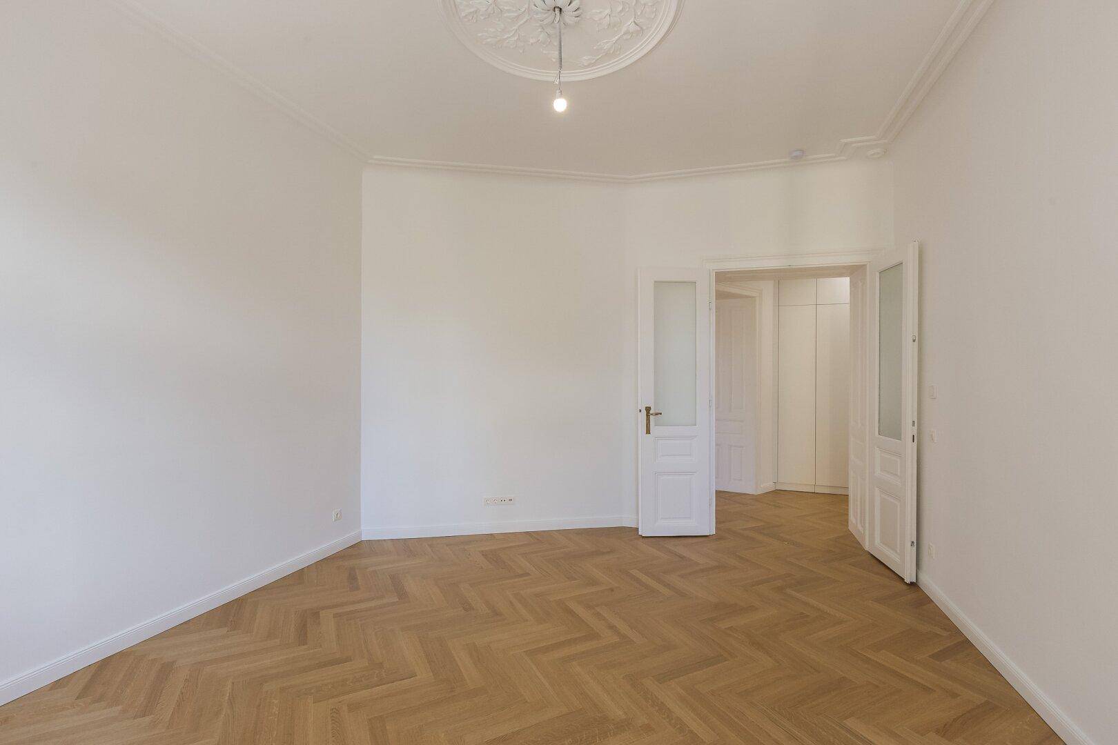 Helles Zimmer mit Fischgrätparkett, Stuckdecke und Zugang zu einem weiteren Raum.
