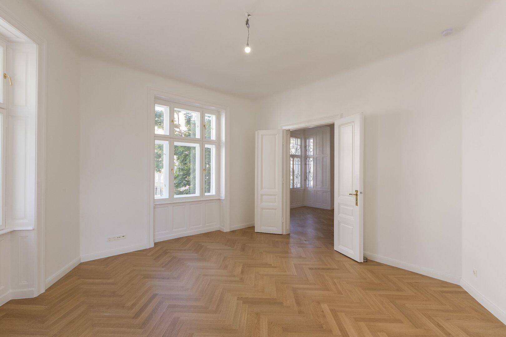 Helles Zimmer mit Fischgrätparkett und Zugang zu einem weiteren Raum durch Flügeltüren.