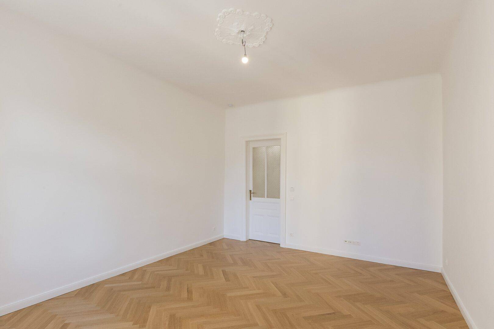 Helles Zimmer mit Fischgrätparkett, Stuckdecke und einer eleganten Tür.