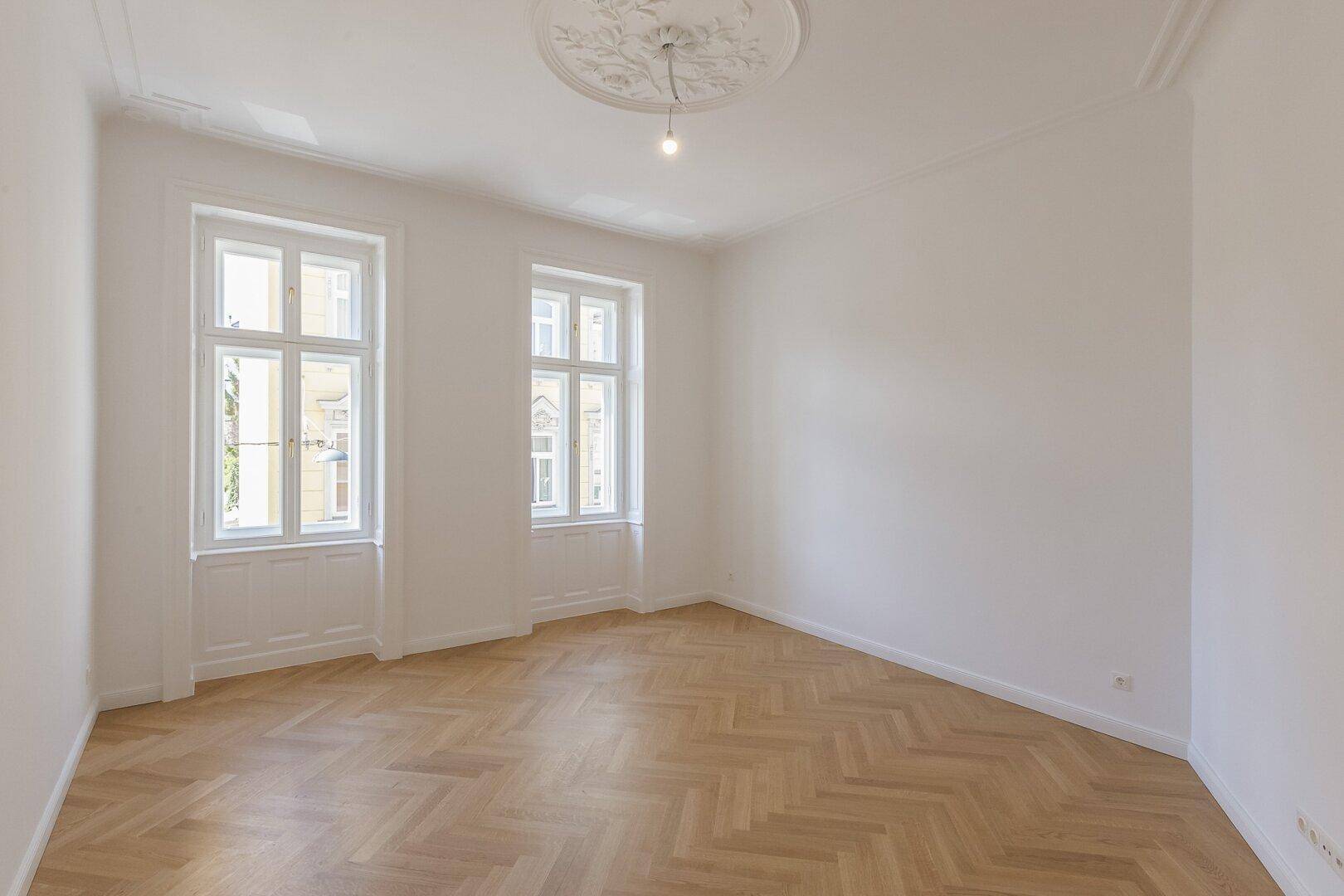 Geräumiges Zimmer mit Fischgrätparkett, Stuckdecke und hohen Fenstern.