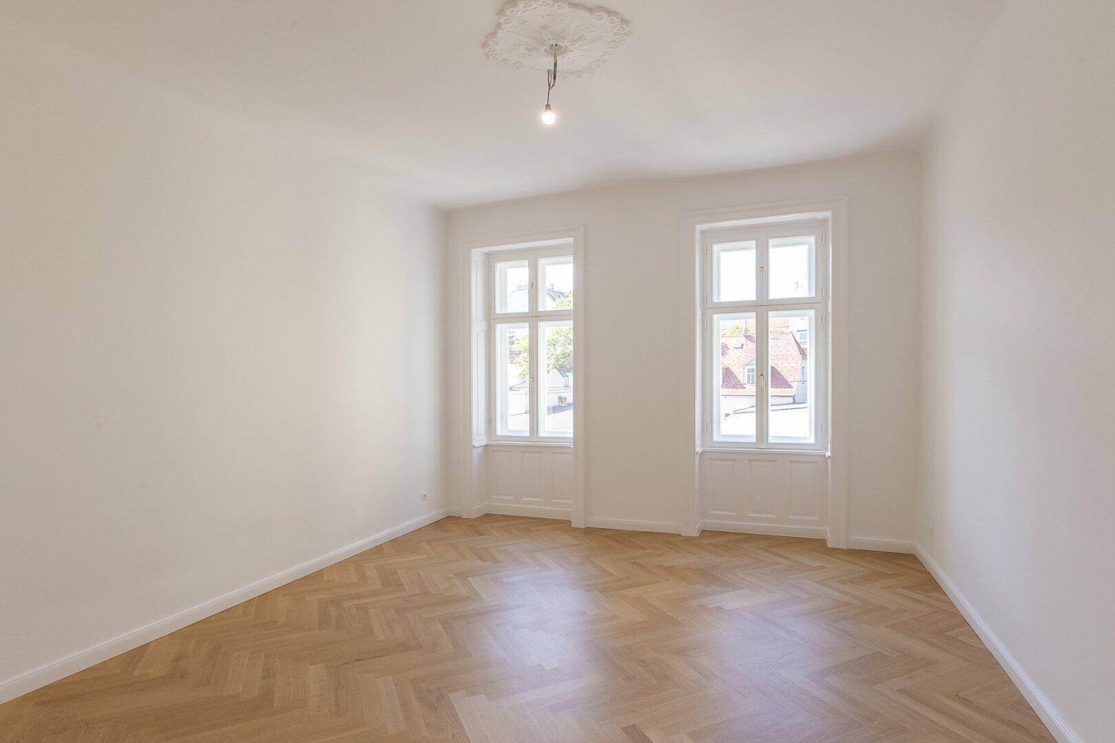 Lichtdurchflutetes Zimmer mit Fischgrätparkett, Stuckdecke und Blick auf die Umgebung.