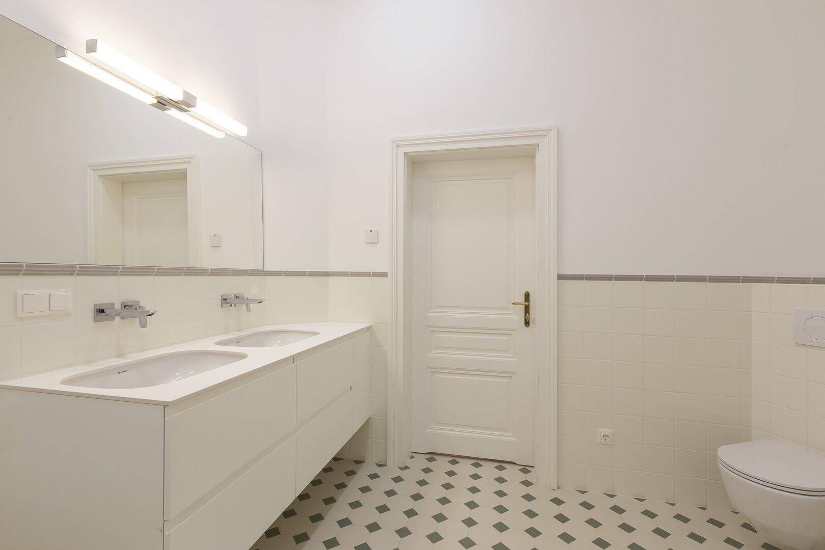 Helles Badezimmer mit Doppelwaschbecken, Toilette und gemusterten Bodenfliesen.