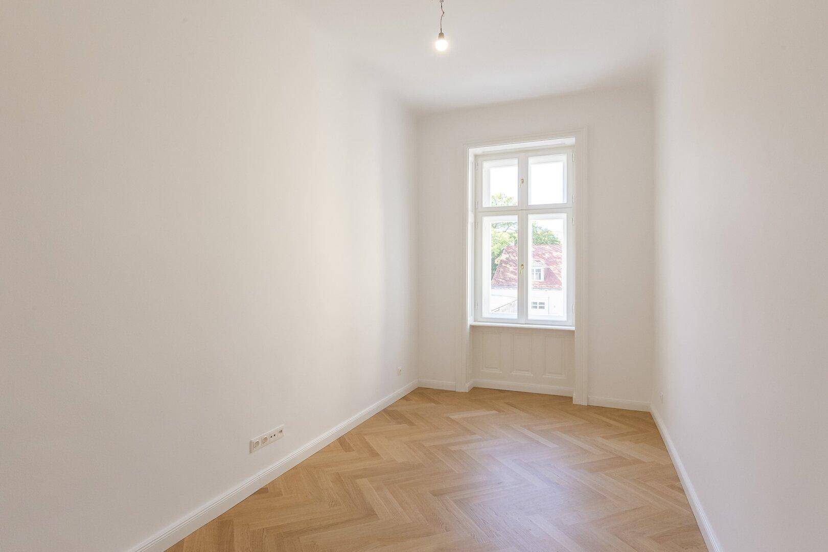 Kleineres Zimmer mit Fischgrätparkett, hohen Decken und einem Fenster.