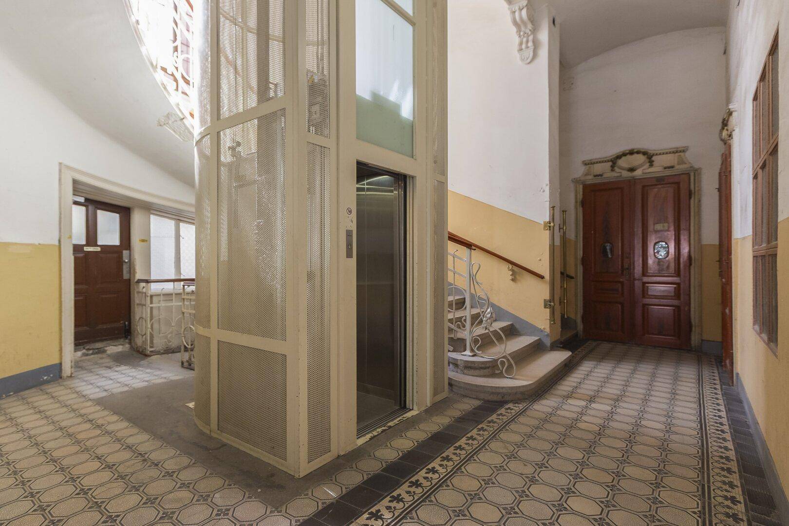 Historischer Hausflur mit originalen Fliesen, Treppenhaus und einem modernen Aufzug.
