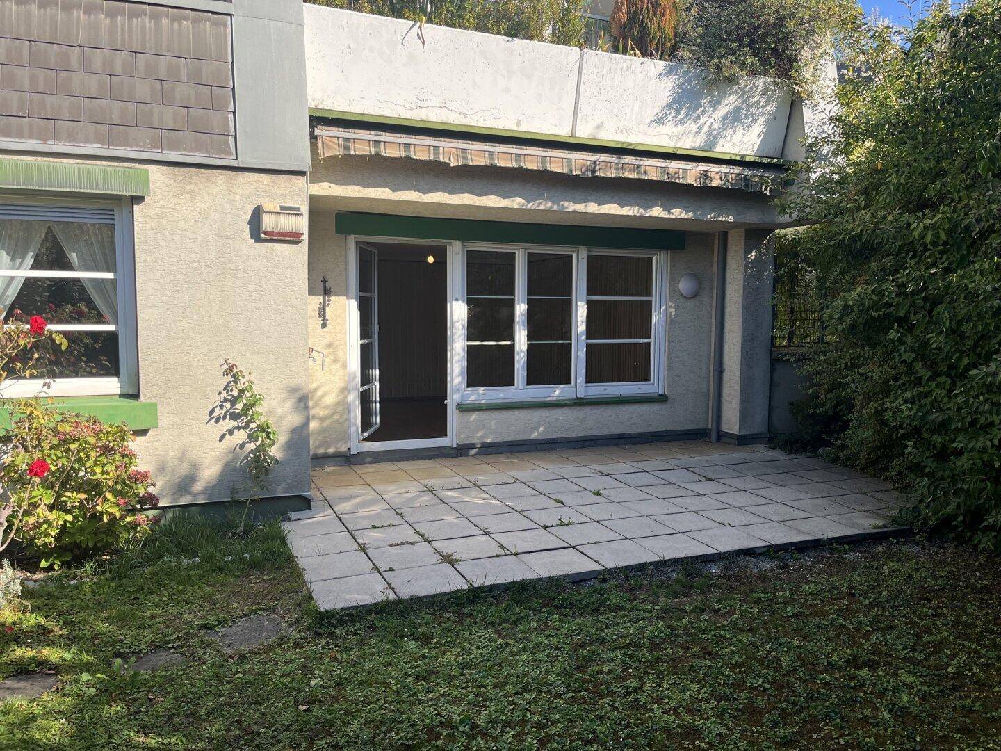 Erdgeschoss-Terrasse mit Markise und Zugang zu einem Innenraum, umgeben von Grün.