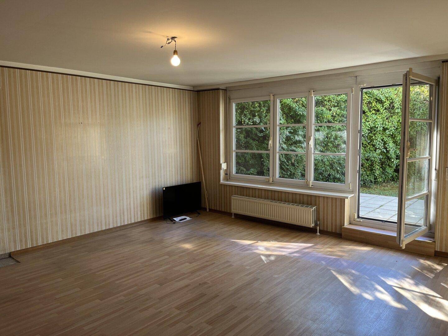 Geräumiges Wohnzimmer mit hellem Holzfußboden und großen Fenstern, die viel Tageslicht hereinlassen.