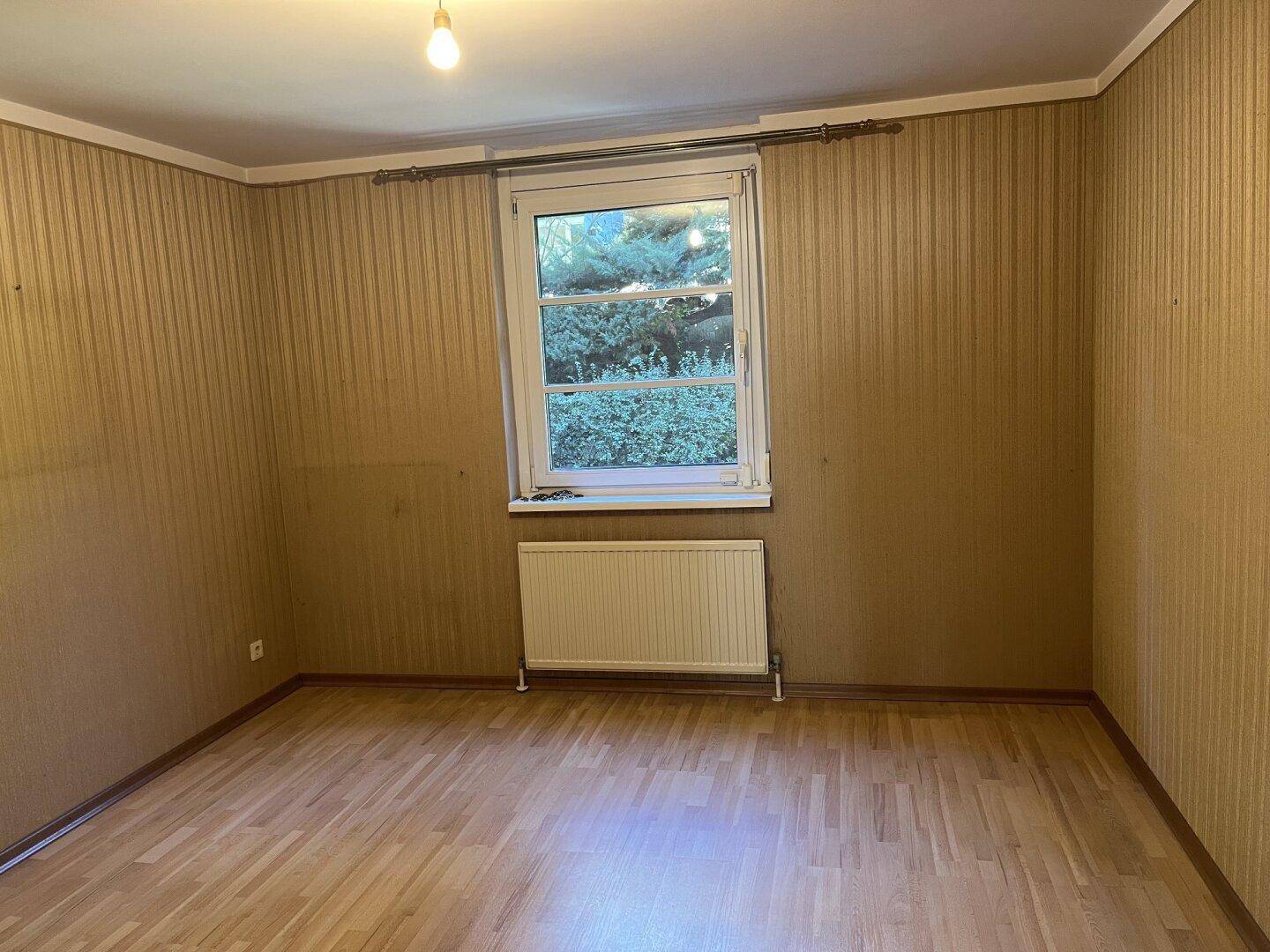 Helles Zimmer mit gestreifter Tapete, Holzfußboden und einem Fenster mit Blick ins Grüne.